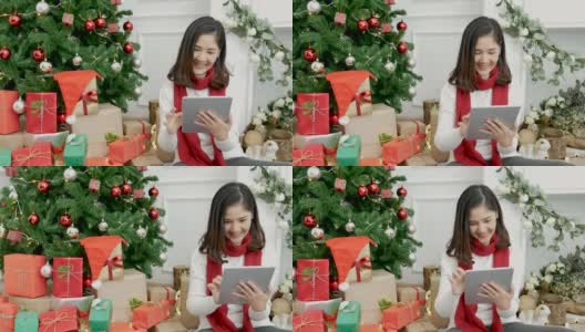 亚洲女人用智能平板电脑坐在圣诞树下的圣诞礼物礼品盒在舒适的客厅。女人手触屏微笑网购，幸福时光圣诞佳节冬季喜庆高清在线视频素材下载