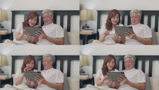 亚洲老年夫妇在家里使用平板电脑。亚洲老爷爷老爷爷，夫妻高兴地醒来后，躺在家里卧室的床上看电影的概念。缓慢的运动。高清在线视频素材下载