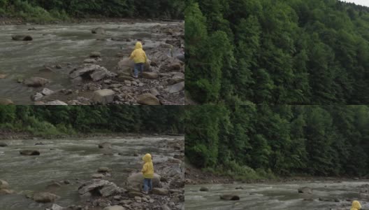 小学前班的孩子在山河附近。穿黄色雨衣的小孩向河里扔石头。男孩从岸上把鹅卵石扔进水里。度假，去露营，春天，野生自然，家庭观念高清在线视频素材下载