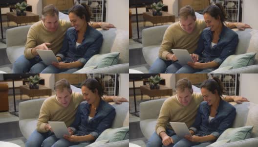 一对幸福的夫妇在家具店看着平板电脑上的产品，坐在沙发上又说又笑高清在线视频素材下载