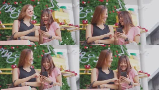 两位快乐的亚洲年轻女性在百货商店的户外购物在销售季节在年底。女孩们拿着购物袋，快乐地走着，微笑着一起买东西高清在线视频素材下载