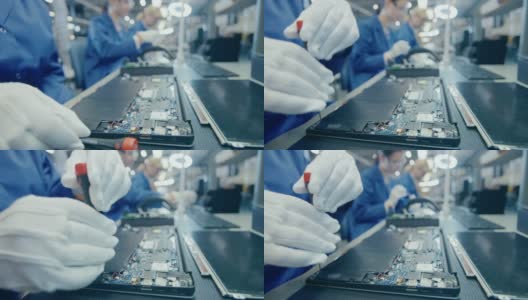 一个穿着蓝色工作服的电子工厂女工用螺丝刀组装笔记本电脑的主板的特写。拥有多名员工的高科技工厂设施。高清在线视频素材下载