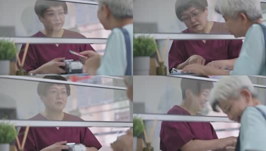 亚裔华人老年女性在诊所用信用卡支付医疗费用，由女护士手持信用卡读卡器高清在线视频素材下载