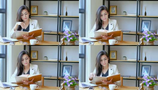 4K视频，美丽的亚洲商务女性坐在电脑笔记本前，阅读和翻动笔记本页与思考的脸在工作空间早上，商务人士的生活方式。高清在线视频素材下载