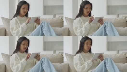 一位亚洲妇女坐在客厅的沙发上使用平板电脑。一个快乐的女人在家里用平板电脑看视频和上网高清在线视频素材下载