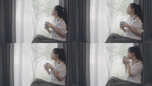 亚洲幸福的女人使用智能手机和喝咖啡在客厅的沙发在家里的背景。以快乐放松为休闲活动理念。4 k慢动作。高清在线视频素材下载