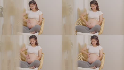 快乐的孕妇坐在床上抱着抚摸着自己的大肚子，年轻的孕妇在家里享受着未来生活的放松，母亲和怀孕的概念，柔软的焦点高清在线视频素材下载