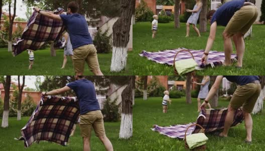 父亲穿着棕色短裤和蓝色衬衫正在铺开格子布准备野餐。在模糊的背景上，快乐的妈妈正在和她的小儿子玩耍。绿色的草。慢动作高清在线视频素材下载