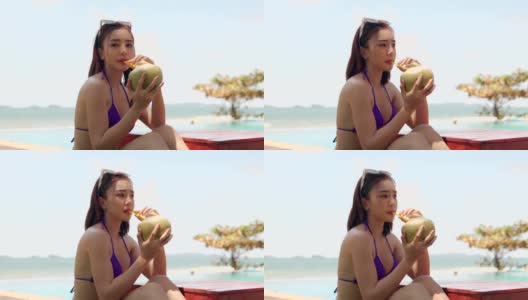 美丽的年轻亚洲女人在紫色比基尼在热带度假泳池喝新鲜的年轻椰子。暑假的概念高清在线视频素材下载