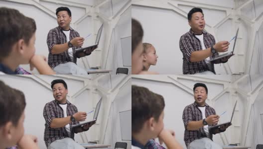 韩国老师坐在桌子上，手里拿着笔记本电脑给学生上课。小学生认真听讲，向老师提出重要问题高清在线视频素材下载