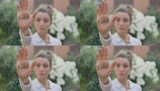 户外女性肖像的年轻成年妇女在白色衬衫显示她的手与口号“我投票了”的鲜花背景，选举日的概念高清在线视频素材下载