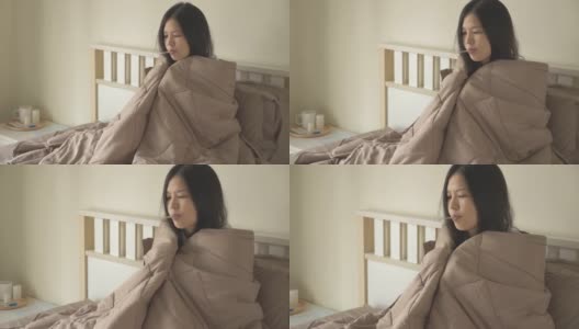 生病的亚洲妇女坐在床上，盖着毯子，有寒冷，颤抖，用温度计测量体温，看着摄像机。高清在线视频素材下载