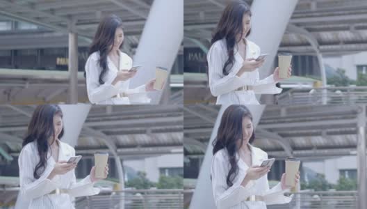 在泰国市中心，年轻的亚洲女商人用着智能手机，面带微笑，边走边喝咖啡。通讯，日常生活技术概念。慢动作高清在线视频素材下载
