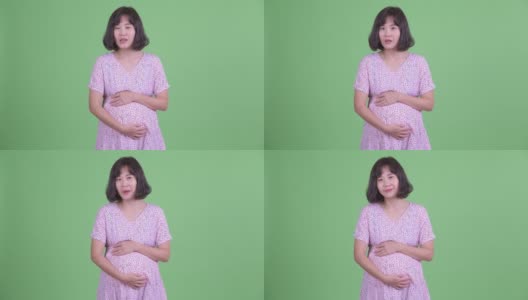 摄影棚拍摄了美丽的亚洲孕妇说话和抱着肚子在绿色的摄影棚背景高清在线视频素材下载