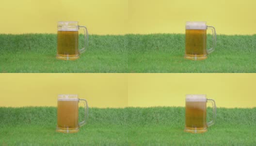 将冰凉爽口的泡沫啤酒倒进大杯子里，杯子立在人造草坪的绿草上，黄色背景，正面观景。小玩具足球滚了出来。高清在线视频素材下载