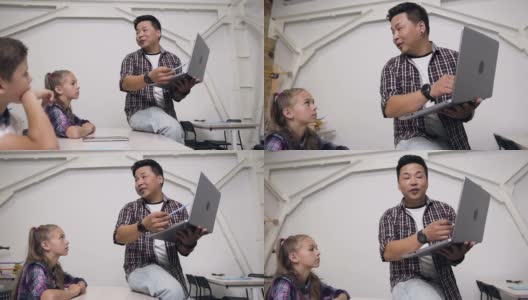 年轻的韩国老师坐在桌子上，手里拿着笔记本电脑给学生上课。小学生认真听讲，向老师提出重要问题高清在线视频素材下载