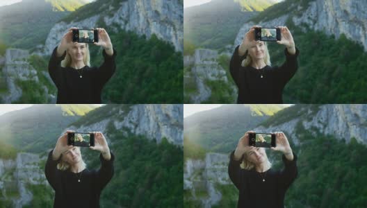 美丽的金发女孩用她的智能手机自拍，拿着屏幕对着相机。在背景风景优美的青山和山景。高清在线视频素材下载