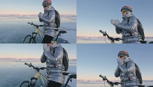 女人在冰上骑自行车。这个女孩穿着银色的羽绒服，背着自行车背包，戴着头盔。女人在用摩擦加热她的手。高清在线视频素材下载