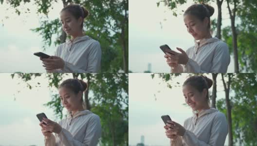 一幅亚洲年轻美女的肖像，在公园的街道上慢跑锻炼后，站在那里看着智能手机，用手机打字与朋友和家人聊天。高清在线视频素材下载