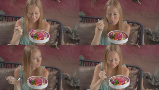 年轻女子坐在美丽的咖啡馆里，享受着盛在碗里的彩色麦片奶昔。健康饮食的概念。Slowmotion拍摄高清在线视频素材下载