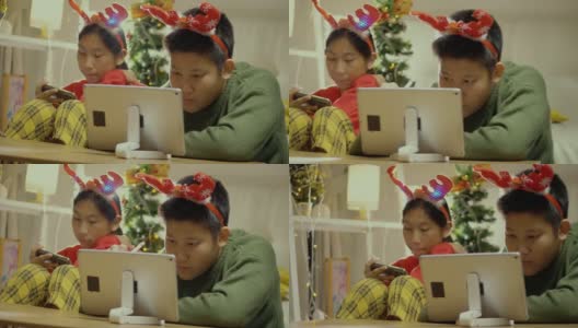 快乐的亚洲儿童穿着毛衣和发带使用智能手机和平板电脑在晚上一起在家里以圣诞装饰为背景，假日概念。高清在线视频素材下载