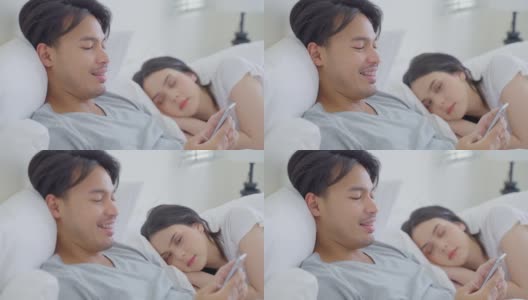 睡在床上的亚洲妻子试图偷看丈夫的手机。迷人有趣的夫妇躺在卧室里，年轻的男性打字短信，而女性女朋友在家里睡觉。高清在线视频素材下载