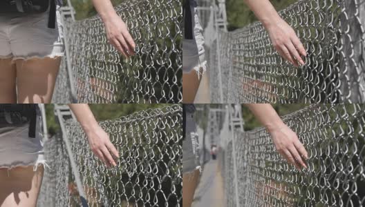 女性的手沿着网格表面移动。年轻女子的手臂碰到金属栅栏。女孩走在暑假里，牵着他的手指沿着桥的守卫。特写慢动作高清在线视频素材下载