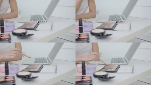 女人的双手在笔记本电脑上学习化妆，用刷子在脸颊上刷在线教程，女性在社交媒体上用调色板和化妆品教学和解释化妆。高清在线视频素材下载