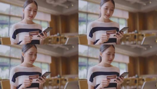 美丽的亚洲年轻女性与开朗的表情，持有智能手机和信用卡，网上银行，快乐的年轻女孩持有塑料信用卡，在咖啡馆使用手机高清在线视频素材下载