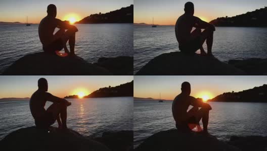 欣赏日落。一个男人坐在岩石沙滩上看风景高清在线视频素材下载