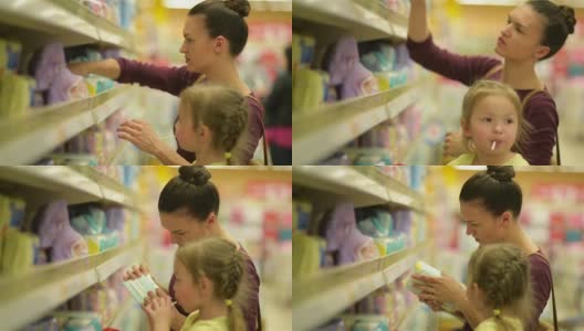 带着棒棒糖的可爱女孩坐在超市手推车里。背景上的母亲选择湿纸巾高清在线视频素材下载