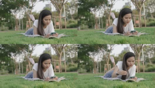 迷人的美丽的年轻亚洲女子放松阅读和使用智能手机，而躺在地板上的草地在一个公园的户外美丽的日落。高清在线视频素材下载