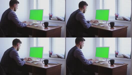 一个年轻人正在电脑上工作，窗户旁边的桌子上有一个绿色屏幕的模型。白人工人在键盘上打字，看着绿色的屏幕高清在线视频素材下载