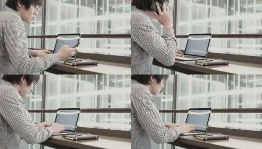 年轻的亚洲商人在工作空间的木桌上使用笔记本电脑和智能手机。男性用手在笔记本电脑键盘上打字。数字时代概念中的自由职业生活方式。高清在线视频素材下载