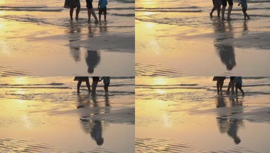 日落时分，在阳光下，一对夫妇带着一个小孩沿着海滩散步，沿着冲浪线。他们的剪影映在水中高清在线视频素材下载