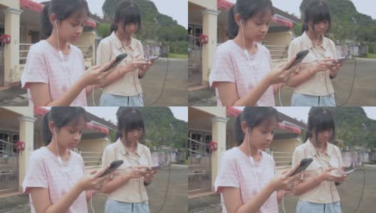 两个可爱的亚洲少女戴着耳机，站在房子外面，喜欢用智能手机聊天和看社交媒体。少女的现代生活。高清在线视频素材下载