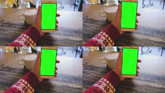 身穿红色圣诞毛衣的女性手持绿色屏幕手机和一杯咖啡放在白色的桌子上。女人使用带有色度键的手机。上网，看内容，轻按中央屏幕高清在线视频素材下载