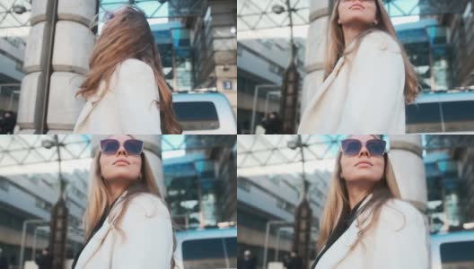 一个戴着眼镜，穿着白色外套的女孩走过城市的商业区。在豪华轿车和公寓的背景下高清在线视频素材下载