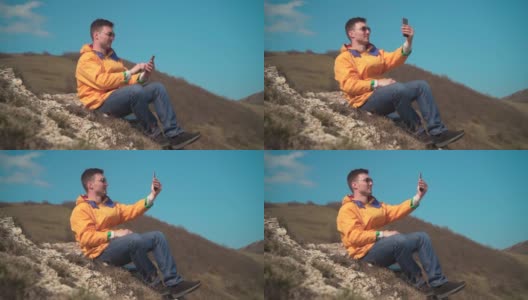 一名身穿黄色夹克、蓝色牛仔裤、戴着眼镜的男子坐在山上，欣赏风景，用手机拍摄视频。高清在线视频素材下载
