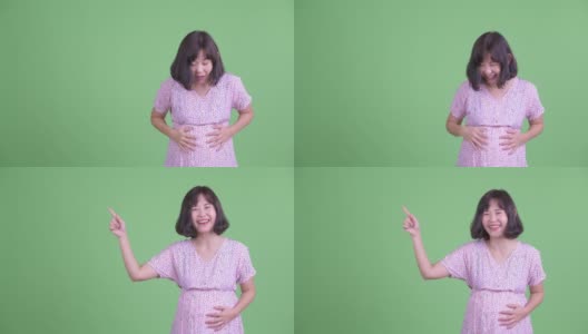 摄影棚拍摄了一个美丽的亚洲孕妇触摸腹部，然后手指着复制空间，在绿色的摄影棚背景下微笑高清在线视频素材下载