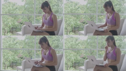 Fit woman使用笔记本电脑，打字，大惊喜，胜利。美丽的亚洲女人在体育。坐在客厅的沙发上。高清在线视频素材下载