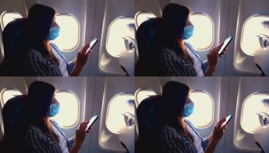 一名年轻女子戴着防护面具，坐在飞机内靠近照明装置的地方使用手机和智能手机。新冠肺炎疫情后恢复航班。开放边界。Post-quarantine旅行高清在线视频素材下载
