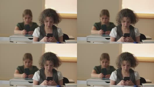 两个女学生坐在学校的课桌前，看着自己的智能手机。儿童和电子产品，青少年和手机在学校高清在线视频素材下载