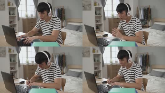 一名亚裔男生戴着耳机，全神贯注地在卧室里写笔记，在笔记本电脑上打字高清在线视频素材下载