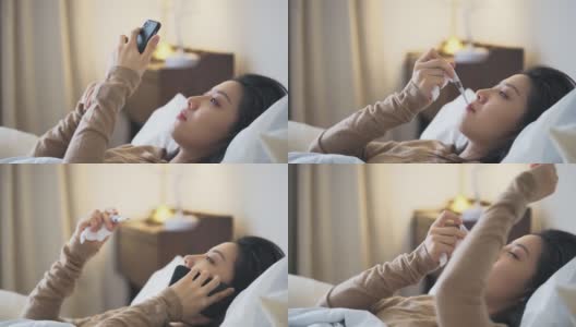 一名亚洲华裔妇女躺在床上，盖着毯子，患感冒和流感，用智能手机和朋友聊天，用数字体温计测量体温高清在线视频素材下载