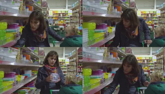一位带着孩子的年轻妇女在超市选购商品。妈妈正用手推车载着一个小女孩。考虑器皿部门的货物。高清在线视频素材下载