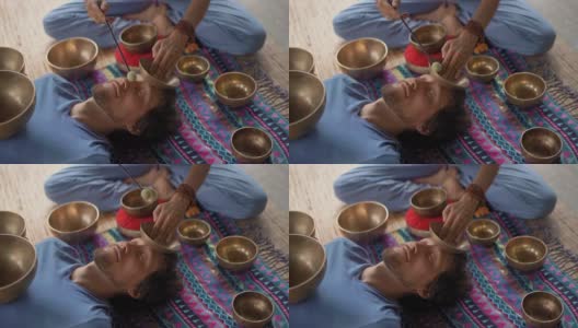 一名精通亚洲圣药的女子为一名年轻男子执行藏碗治疗仪式的超慢镜头。用西藏唱碗冥想。他们在一个凉亭里冥想，背景是美丽的瀑布高清在线视频素材下载