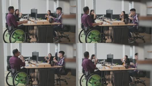 亚洲同事和坐在轮椅上的印度同事在办公室的工作站上分享面包点心高清在线视频素材下载