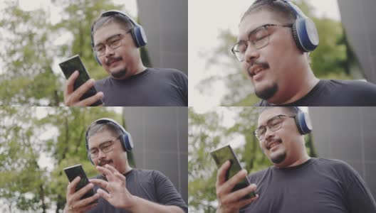 肖像迷人的亚洲男人肥胖，戴着眼镜和胡子微笑自信享受。用智能手机听耳机里的音乐。看着相机，很开心。积极的思考。高清在线视频素材下载