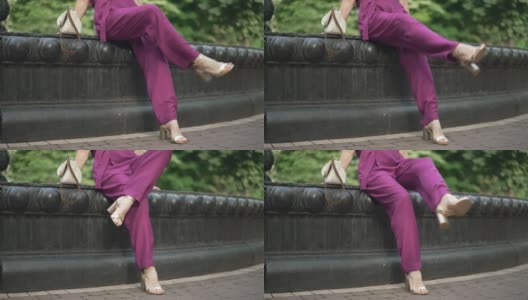 一位无法辨认的女子坐在城市公园的喷泉旁，双腿交叉在膝盖上，慢动作。苗条时尚的白种人女士穿着紫色连衣裤和高跟鞋在夏日户外休息高清在线视频素材下载